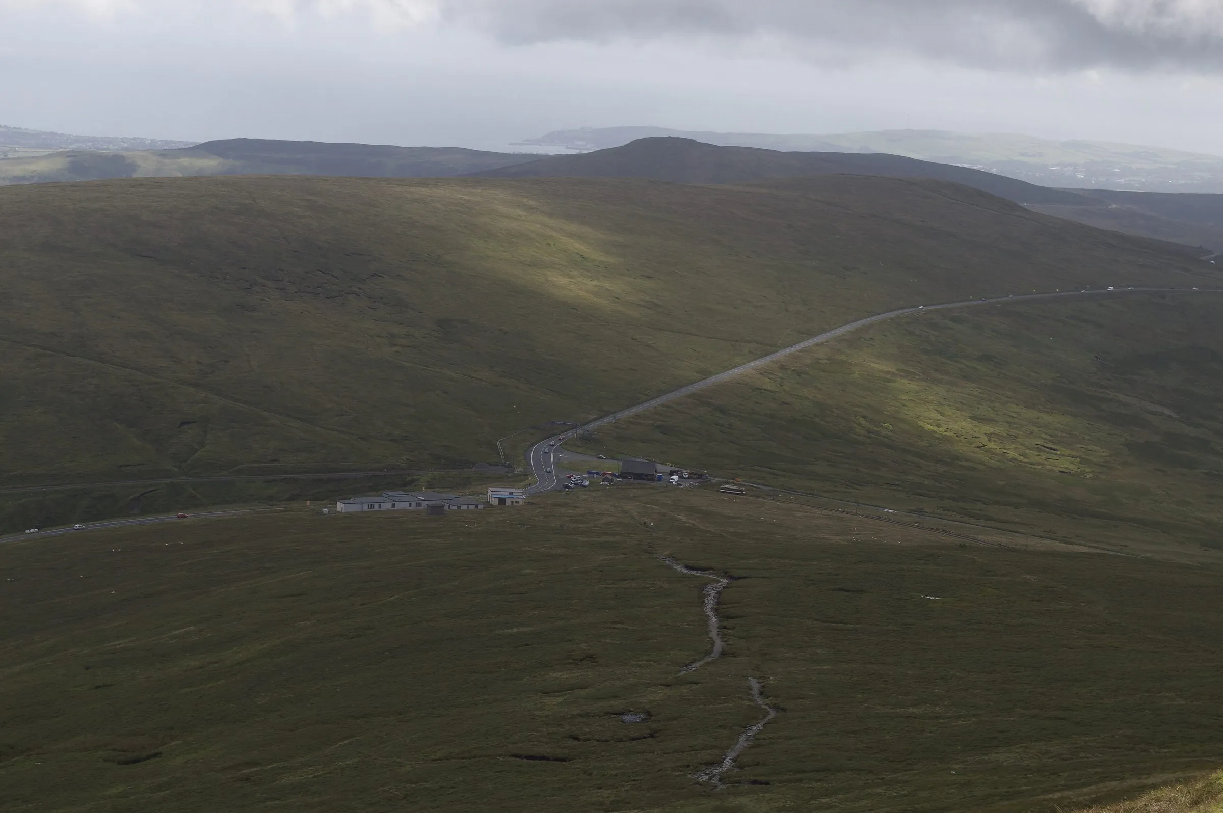 Looking down from Snaefell's slopes to the Bungalow station and the Mountain Road winding across brown moorland, with the sea and hills beyond