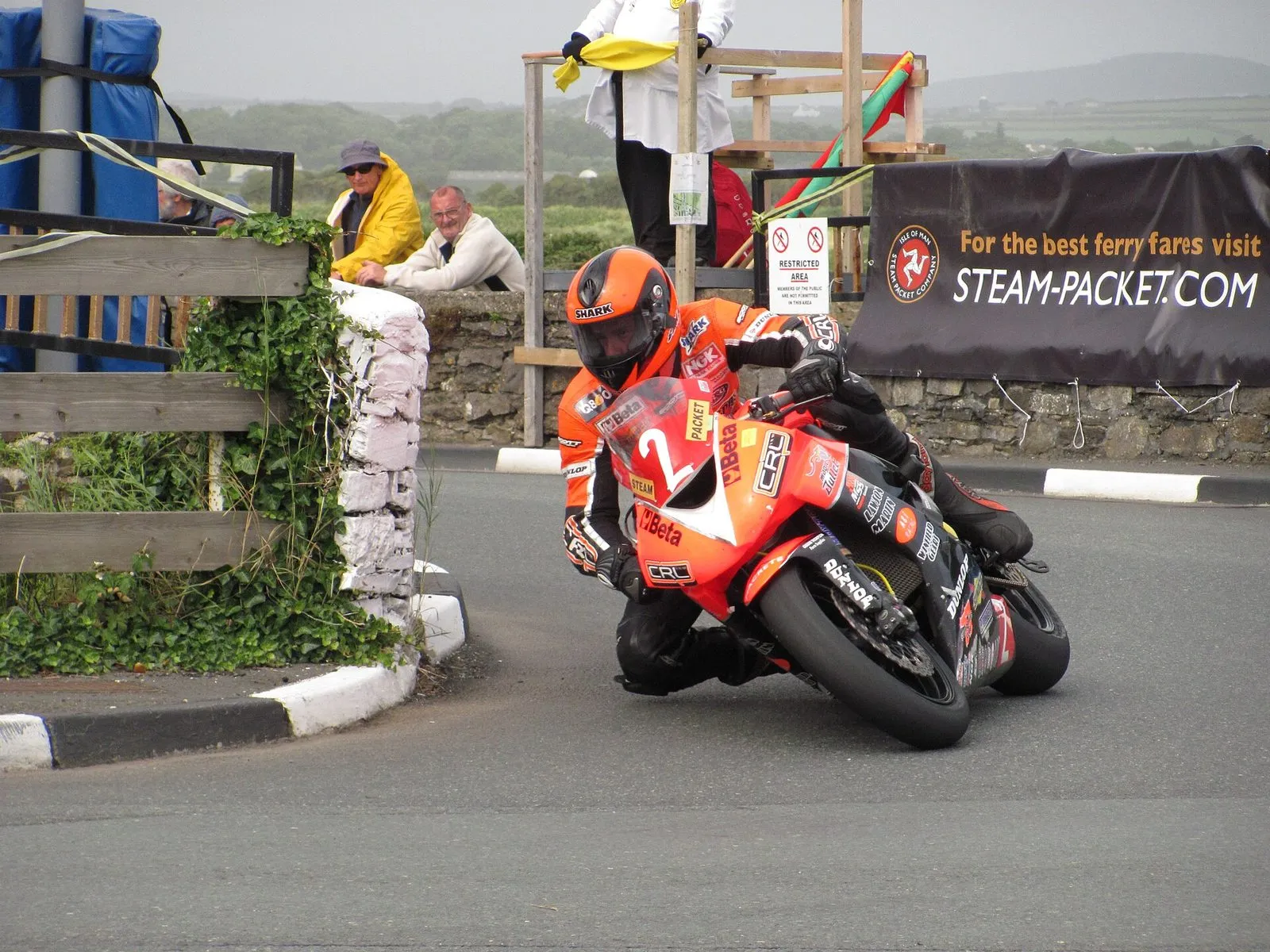 A rider in orange leathers on an orange bike number 2 cornering tightly past a whitewashed wall, with a marshal holding a flag and spectators behind