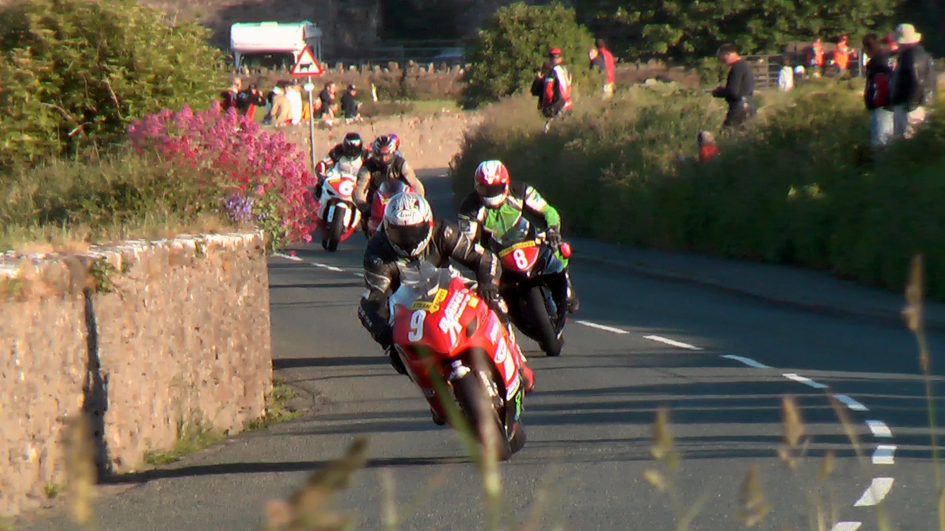 A pack of solo racing bikes led by a red number 9 coming out of a bend between a stone wall and hedges, with spectators on the bank behind