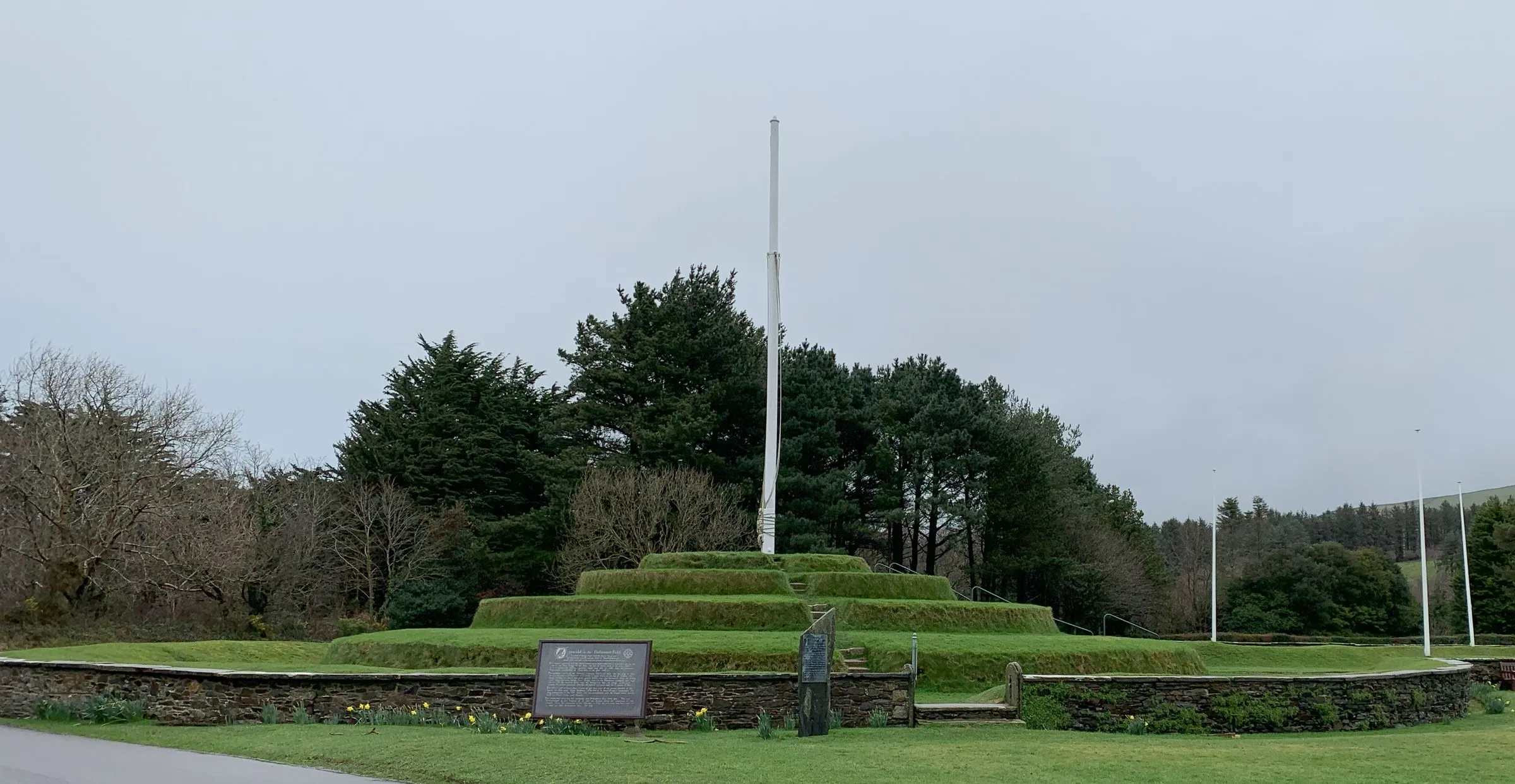 The grassy stepped tiers of Tynwald Hill with a flagpole on top, trees behind and the low wall around its base