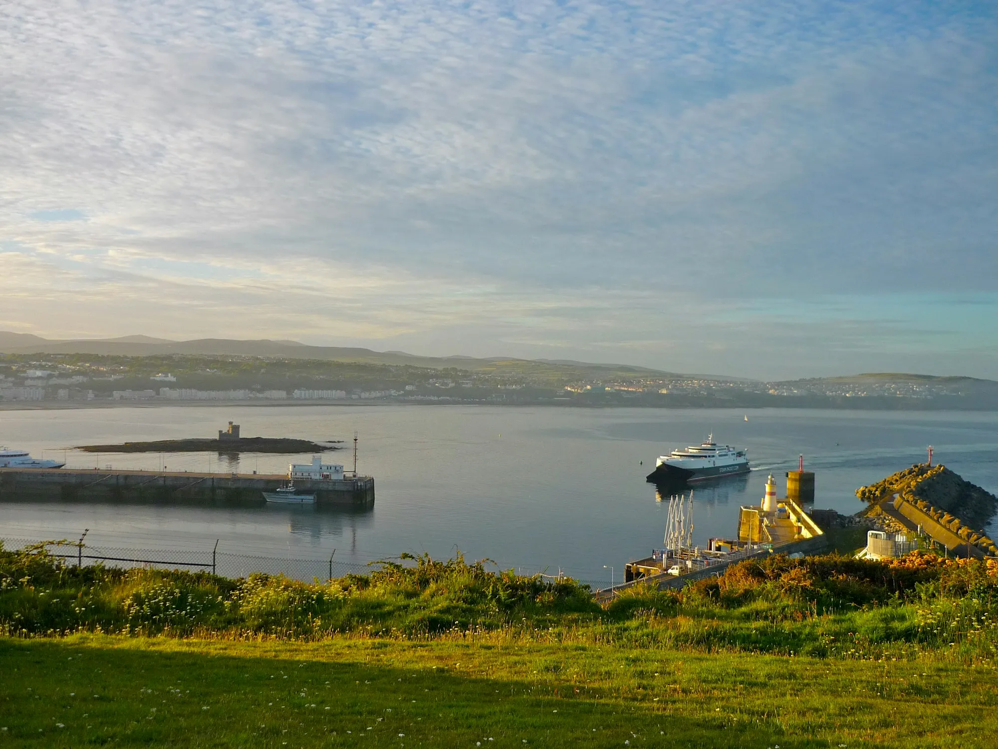 A Steam Packet ferry entering Douglas Harbour in early morning light, seen from the grass on Douglas Head, with Douglas Bay and the hills behind