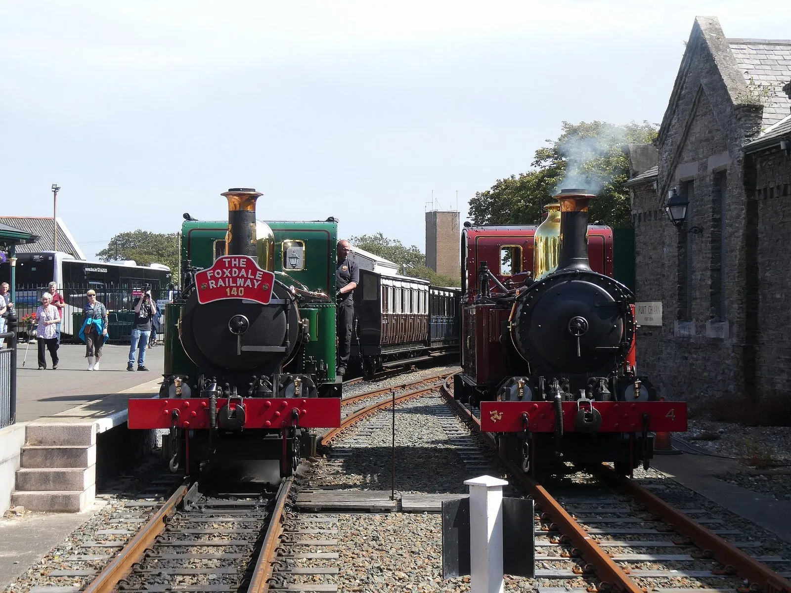 Two steam tank engines, one green and one red, side by side on the tracks at Port Erin station beside a stone building