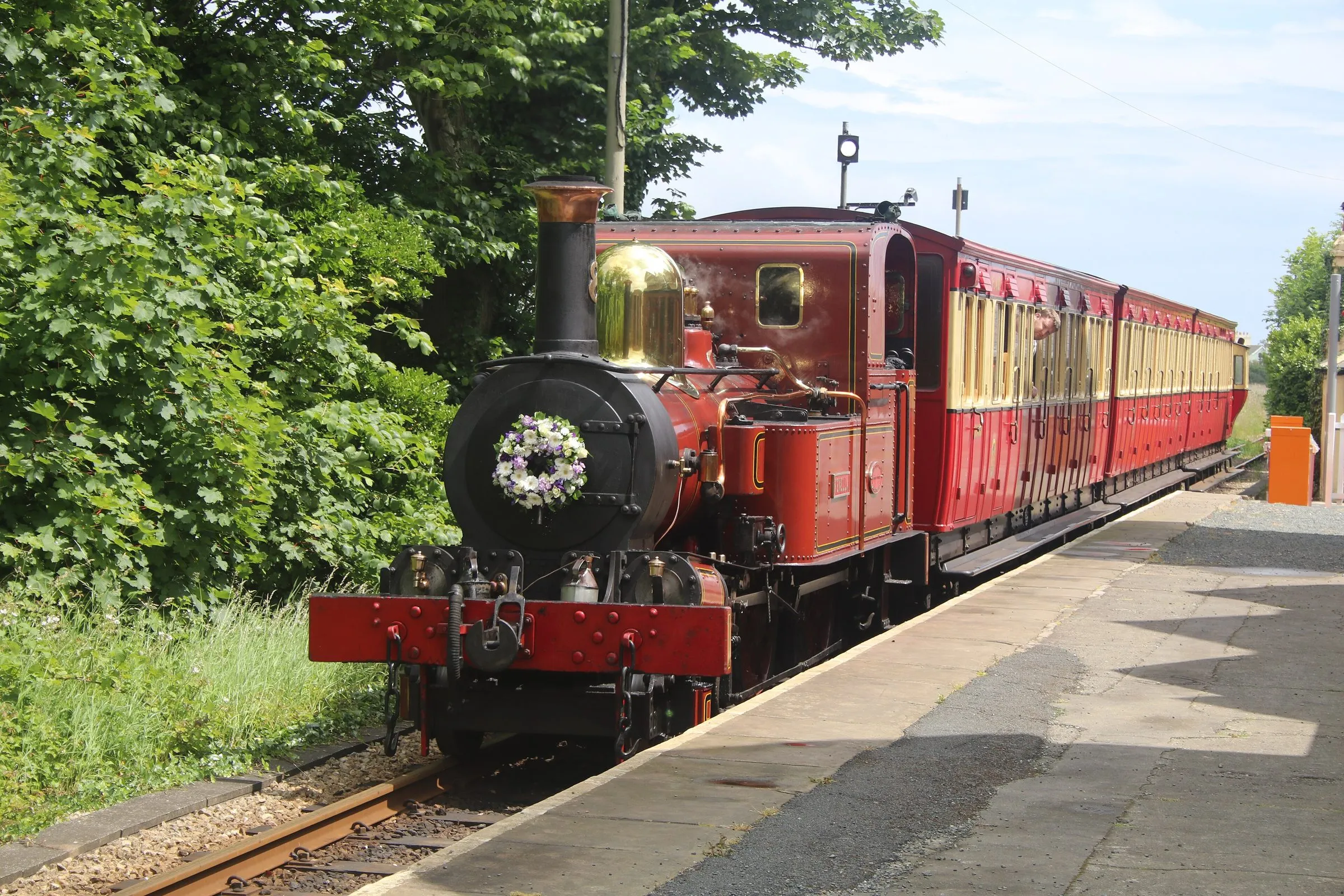 A red Victorian tank engine with a polished brass dome pulling red and cream carriages into Port St Mary station past green trees