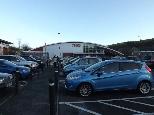 The front of the Tesco superstore on Lake Road, Douglas, with a curved roof and red Tesco sign behind a full car park