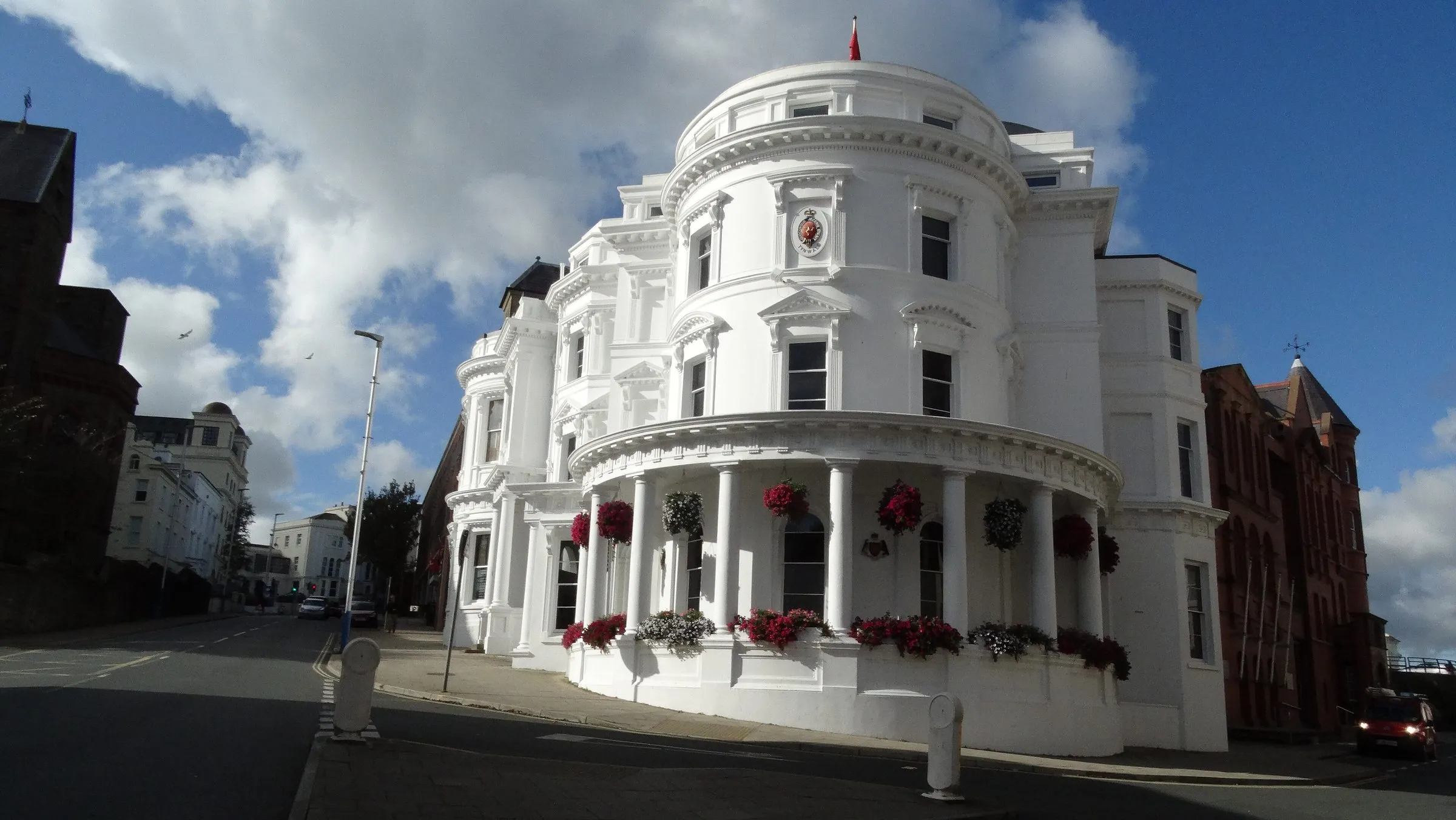 The white, round-fronted Isle of Man Government building on Prospect Hill in Douglas, with hanging baskets under a blue sky