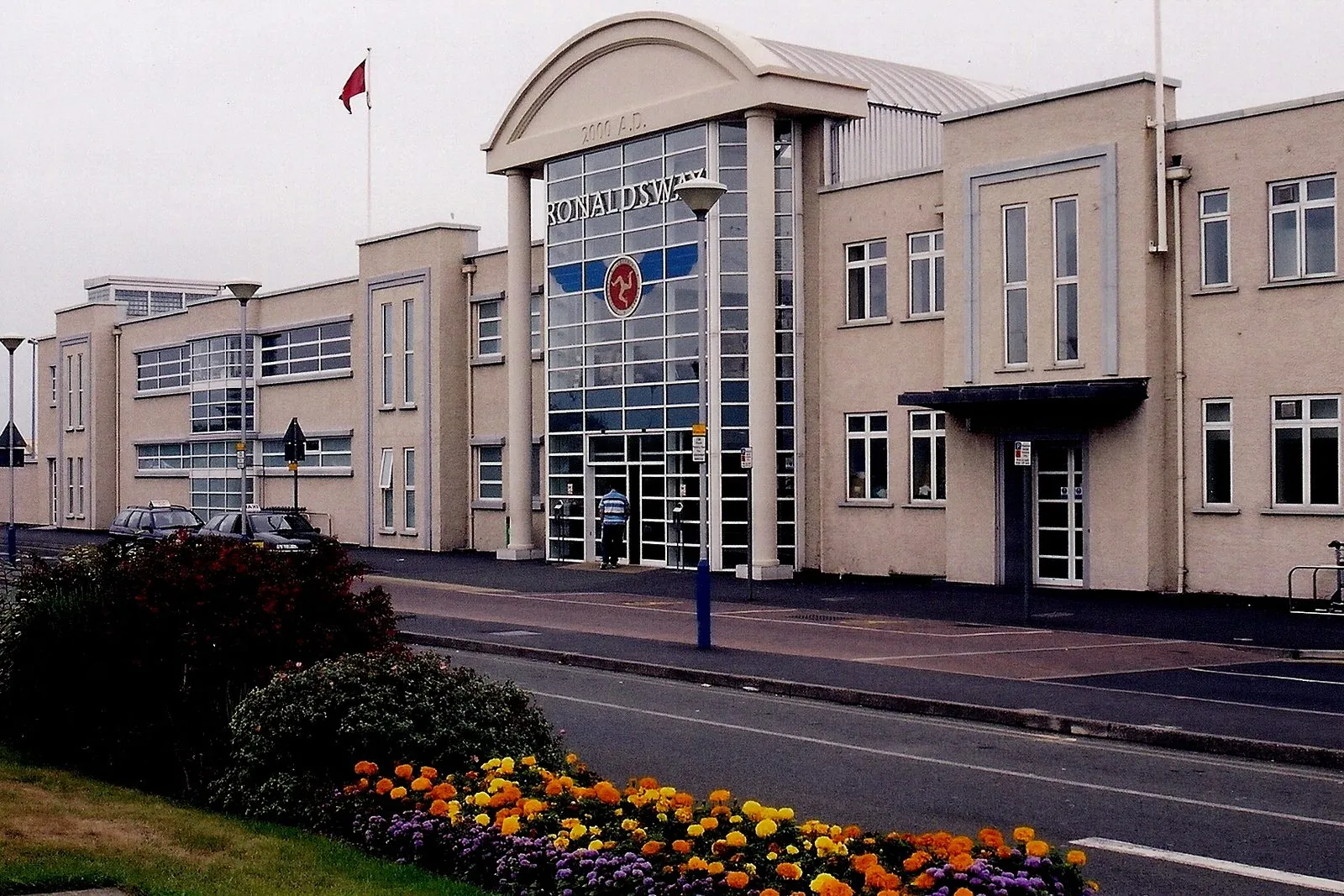 The Ronaldsway Airport terminal building with its glass entrance, cars waiting at the kerb and a bed of orange flowers in front