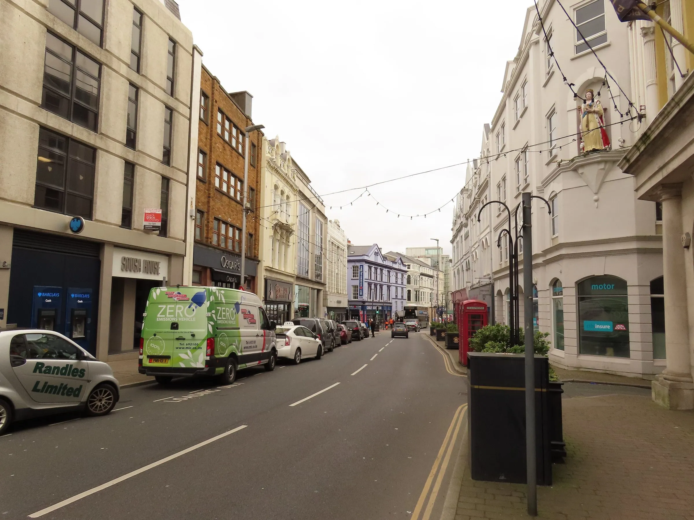 Victoria Street in Douglas city centre, with cars parked along the kerb, shop fronts on both sides and a bus in the distance