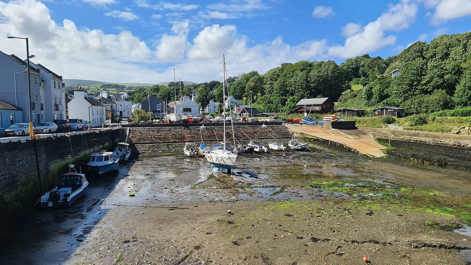 Small boats and a yacht sitting on the sand and weed of Laxey harbour at low tide, with the harbour wall, a slipway and wooded hillside behind