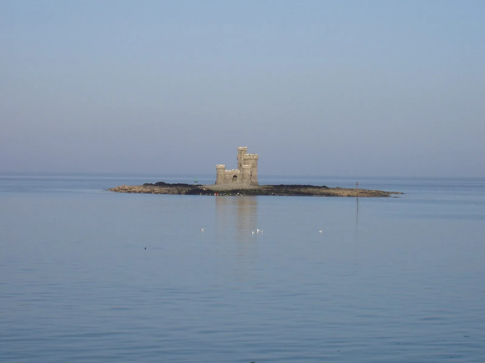 The small castle-like Tower of Refuge on the low, dark rocks of Conister Rock, surrounded by calm blue water in Douglas Bay