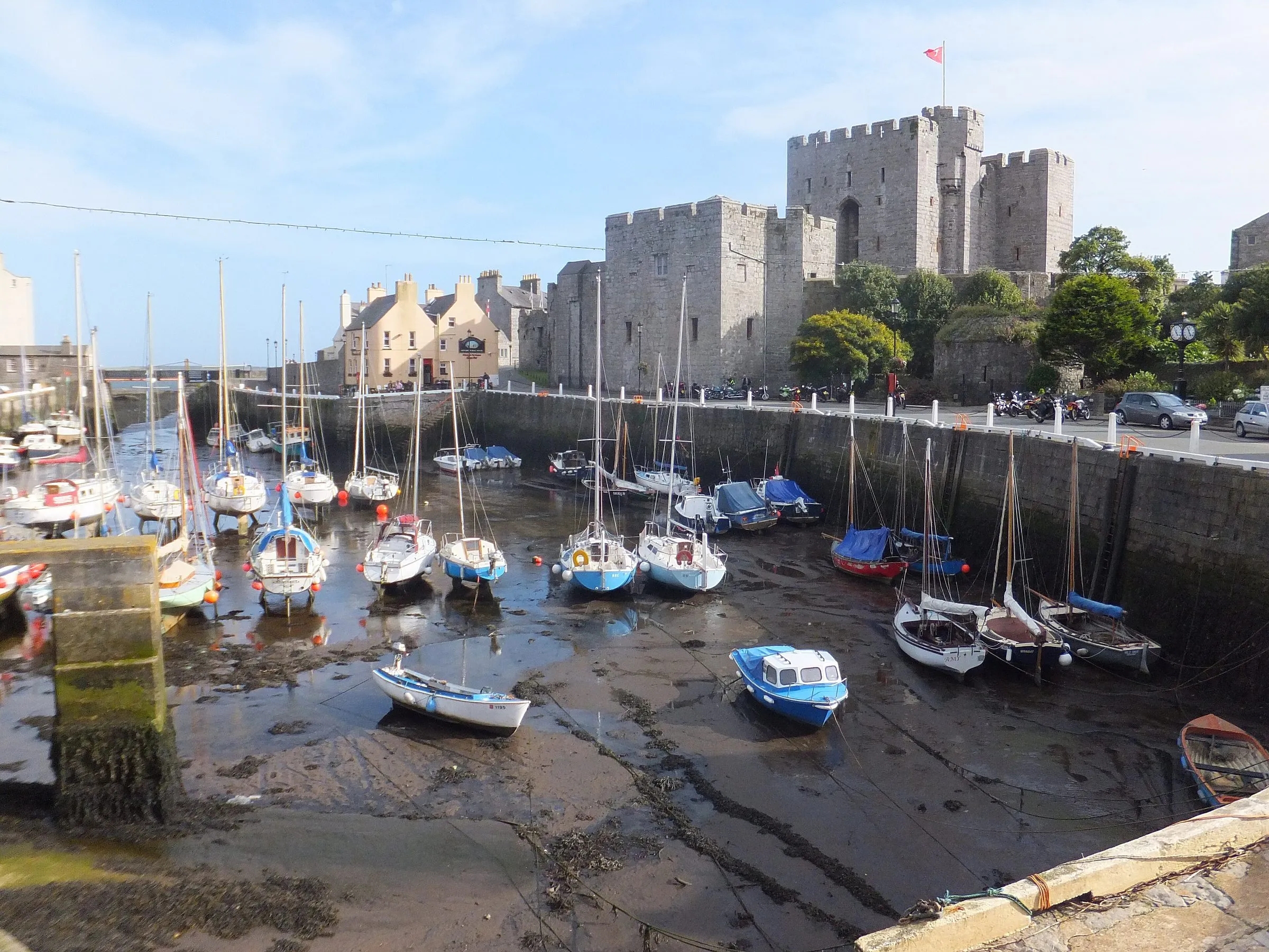 Small yachts and boats sitting on the mud and seaweed of Castletown harbour at low tide, with the grey walls of Castle Rushen behind