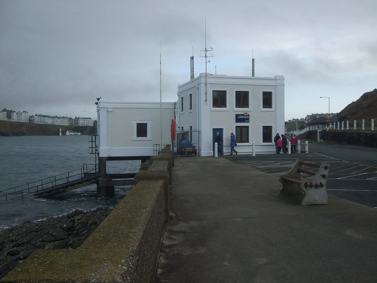 The white two-storey Port Erin lifeboat station on the breakwater, with a slipway down to the sea and the town’s hillside houses behind under a grey sky
