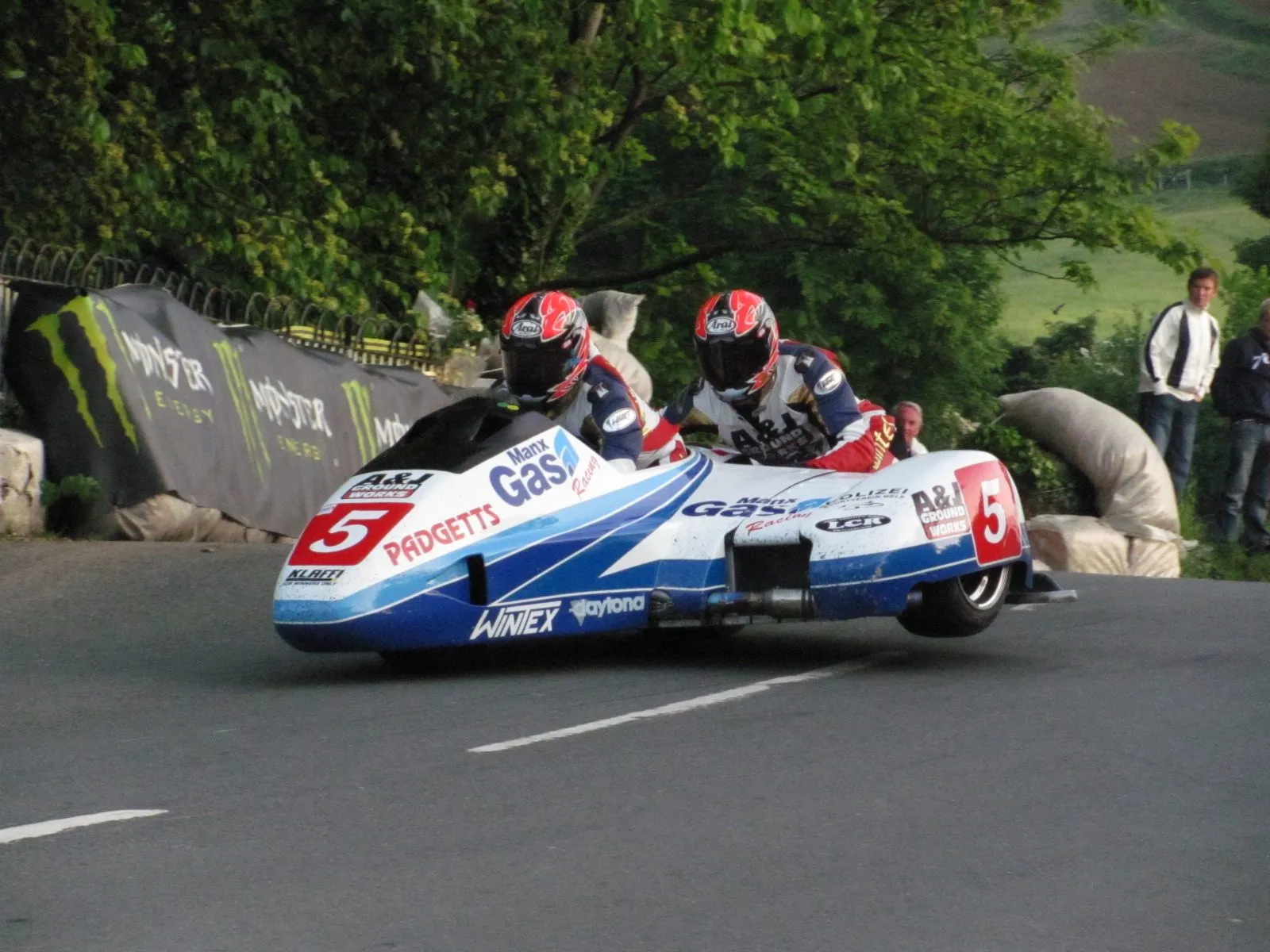 A TT sidecar outfit, number 5, leaning through the bend at Ballaugh Bridge with a driver and passenger in red helmets, and spectators behind the straw bales