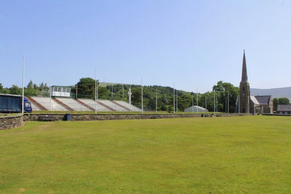 A green field in front of empty grandstands with a tall-spired stone church on the right, and flagpoles lining a path