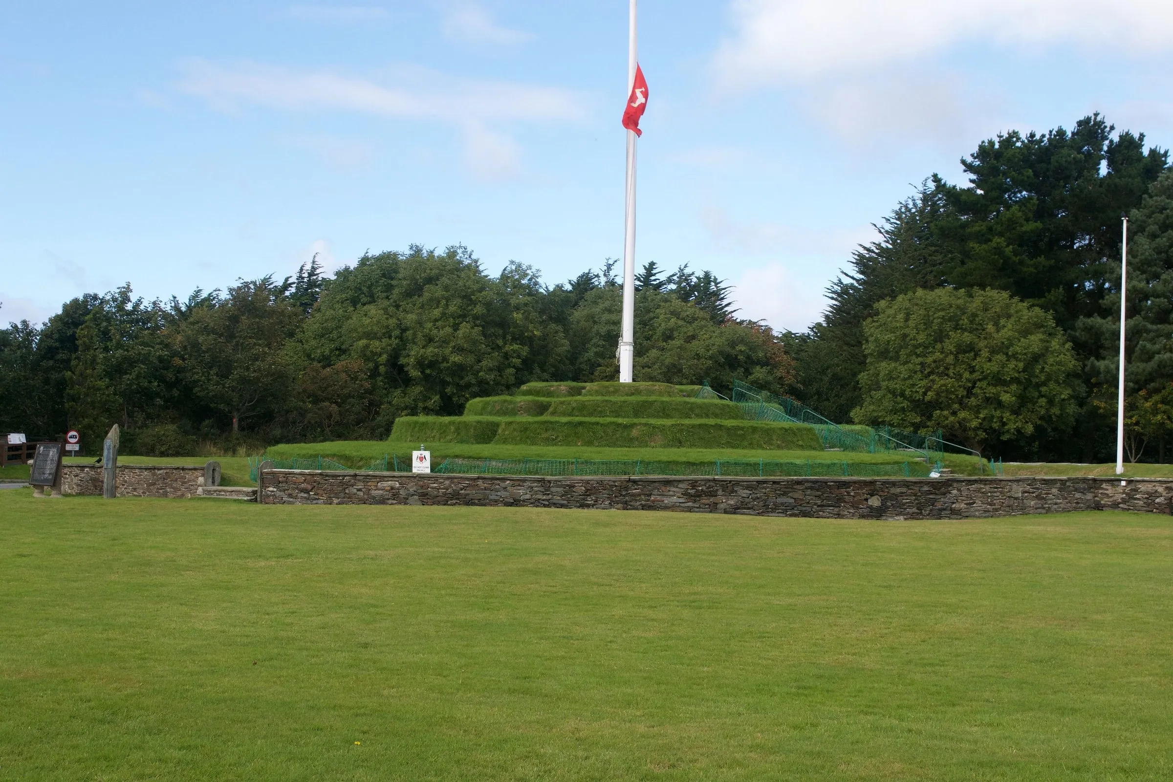 Tynwald Hill at St Johns, a stepped grass mound with a tall flagpole flying the Manx flag, ringed by a low stone wall and trees