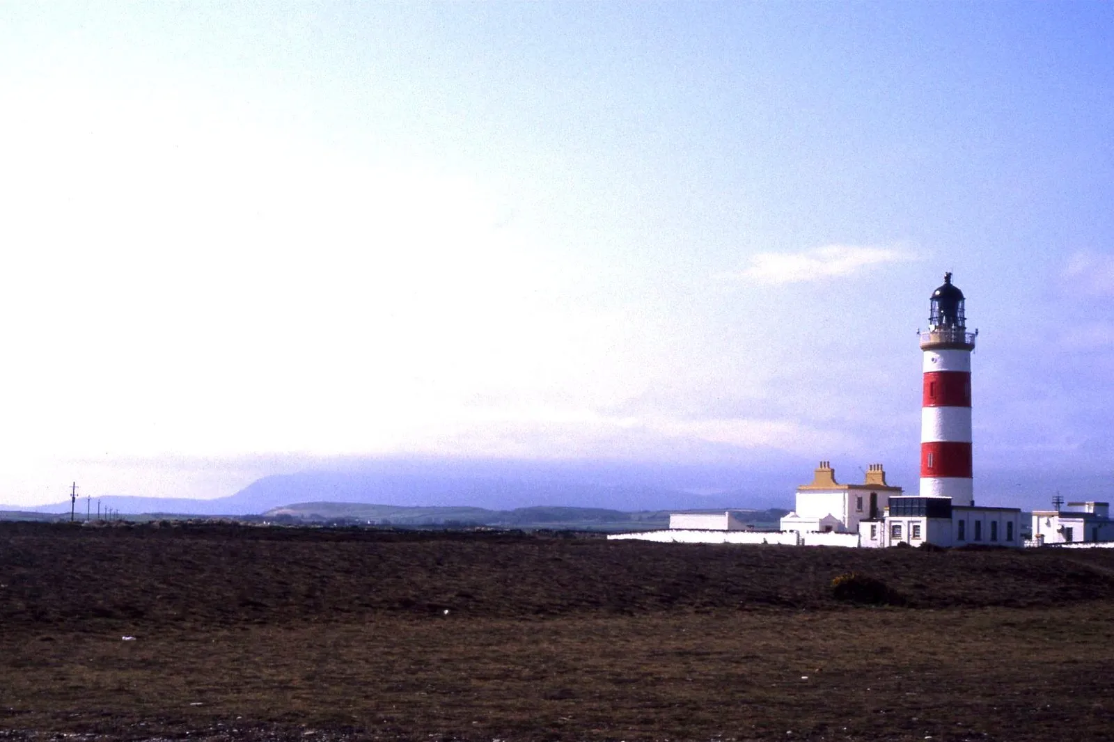 The red-and-white banded Point of Ayre lighthouse beside white keepers’ buildings on a flat, stony plain, with hills on the horizon