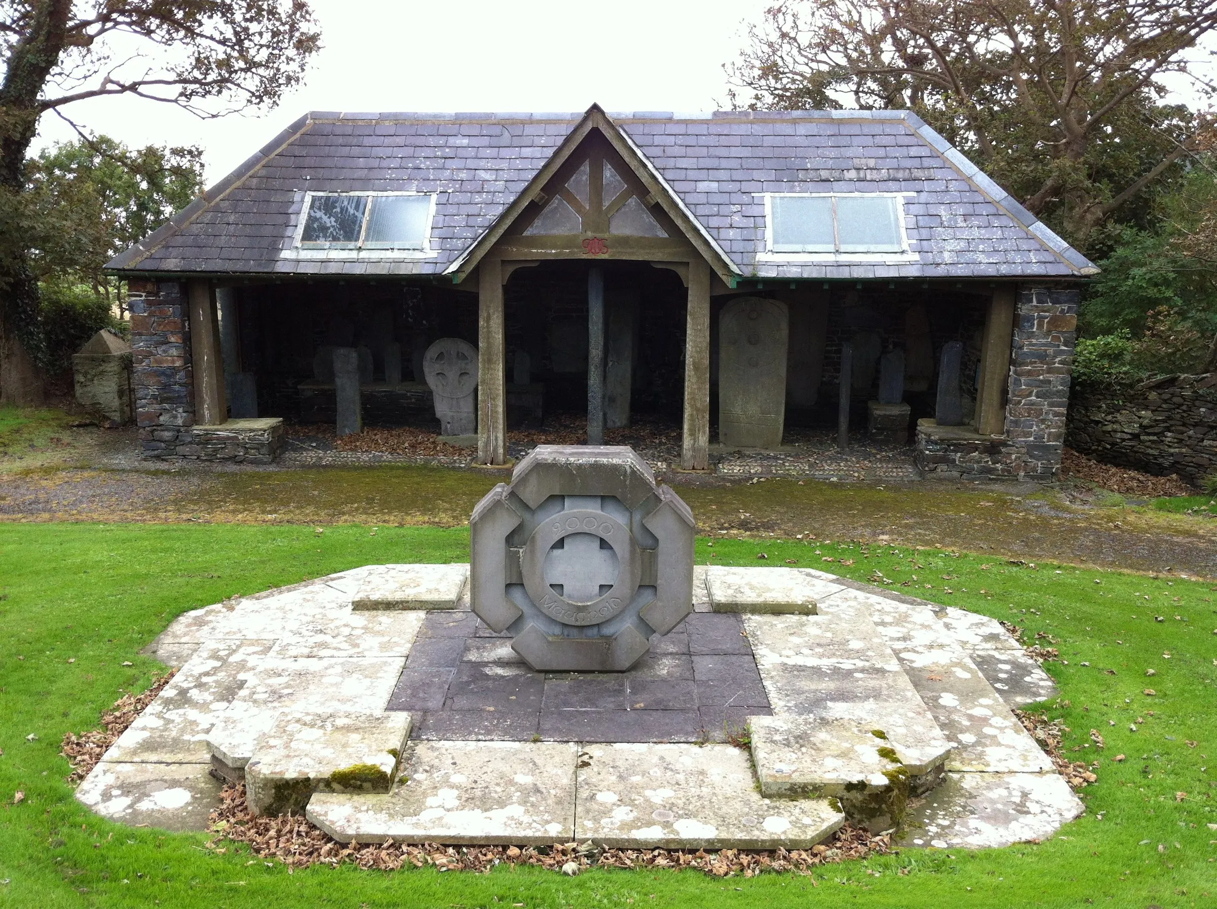 The open-fronted stone cross shelter in Kirk Maughold churchyard, with carved Manx crosses standing inside and a stone memorial on the lawn in front