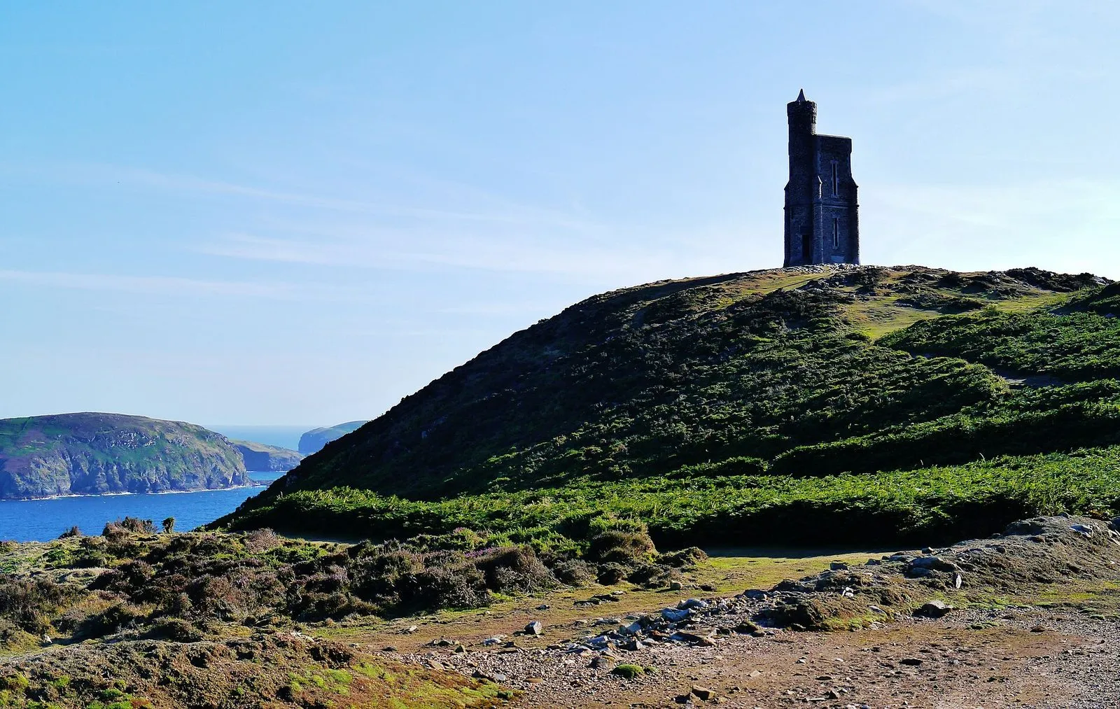 A stone tower on a heather-covered headland under a blue sky, with a green island and the sea in the distance