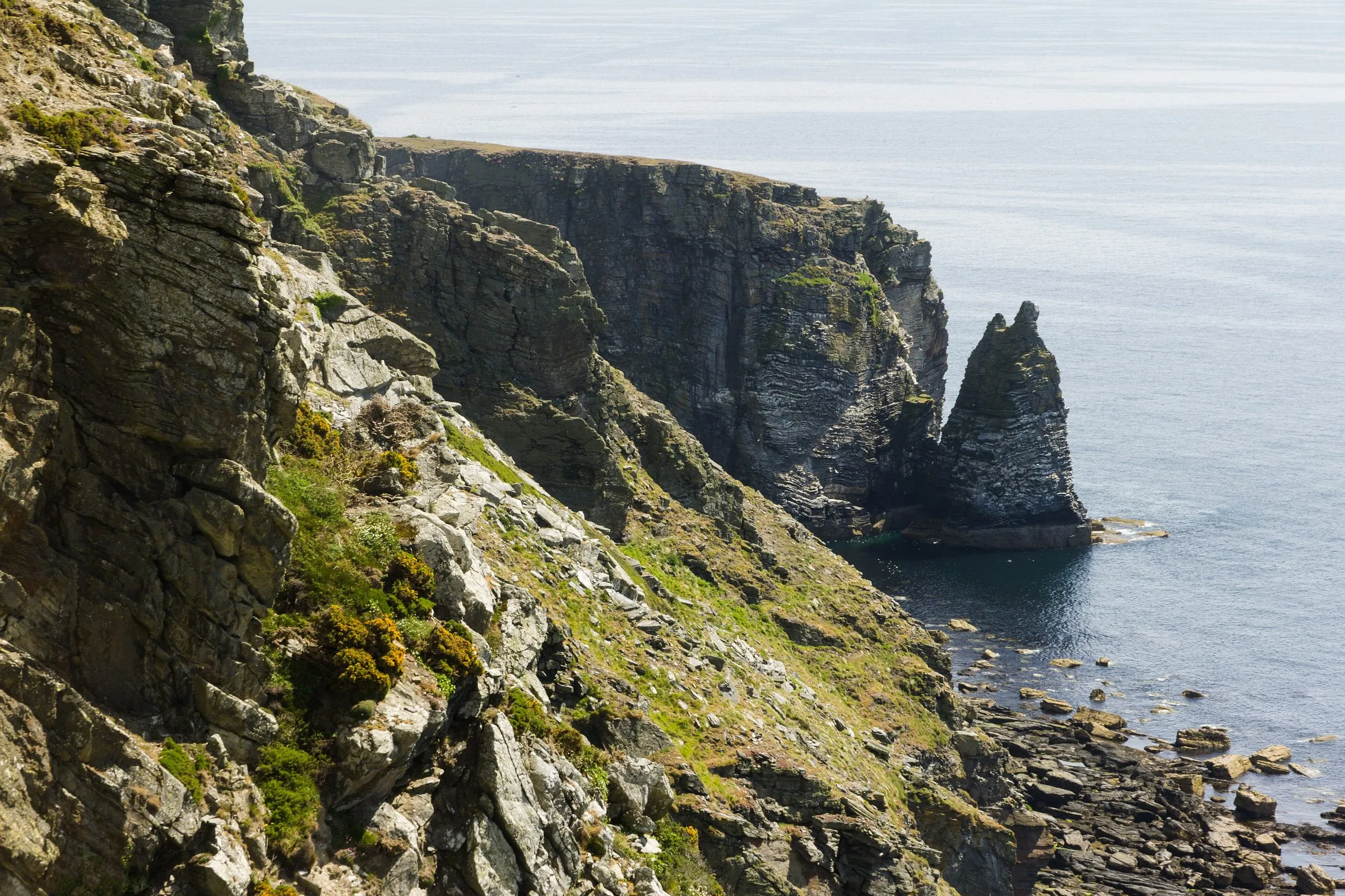 Layered grey sea cliffs and a tall rock stack at the Chasms in the far south of the Isle of Man, with a calm sea below