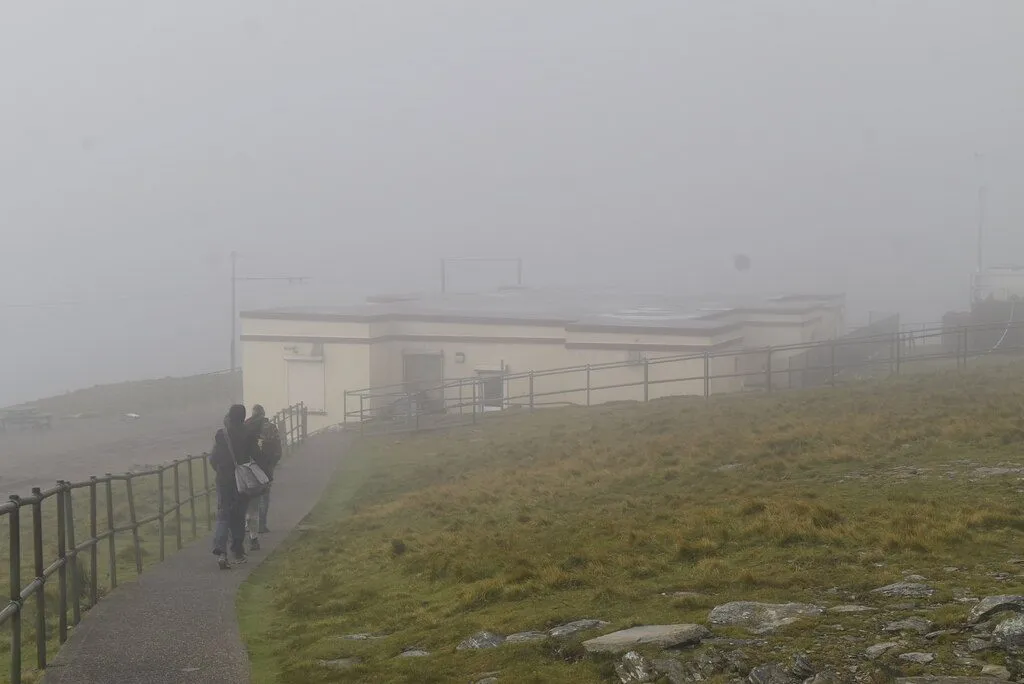 A white single-storey café building on the summit of Snaefell, barely visible in thick fog, with two walkers on the path towards it