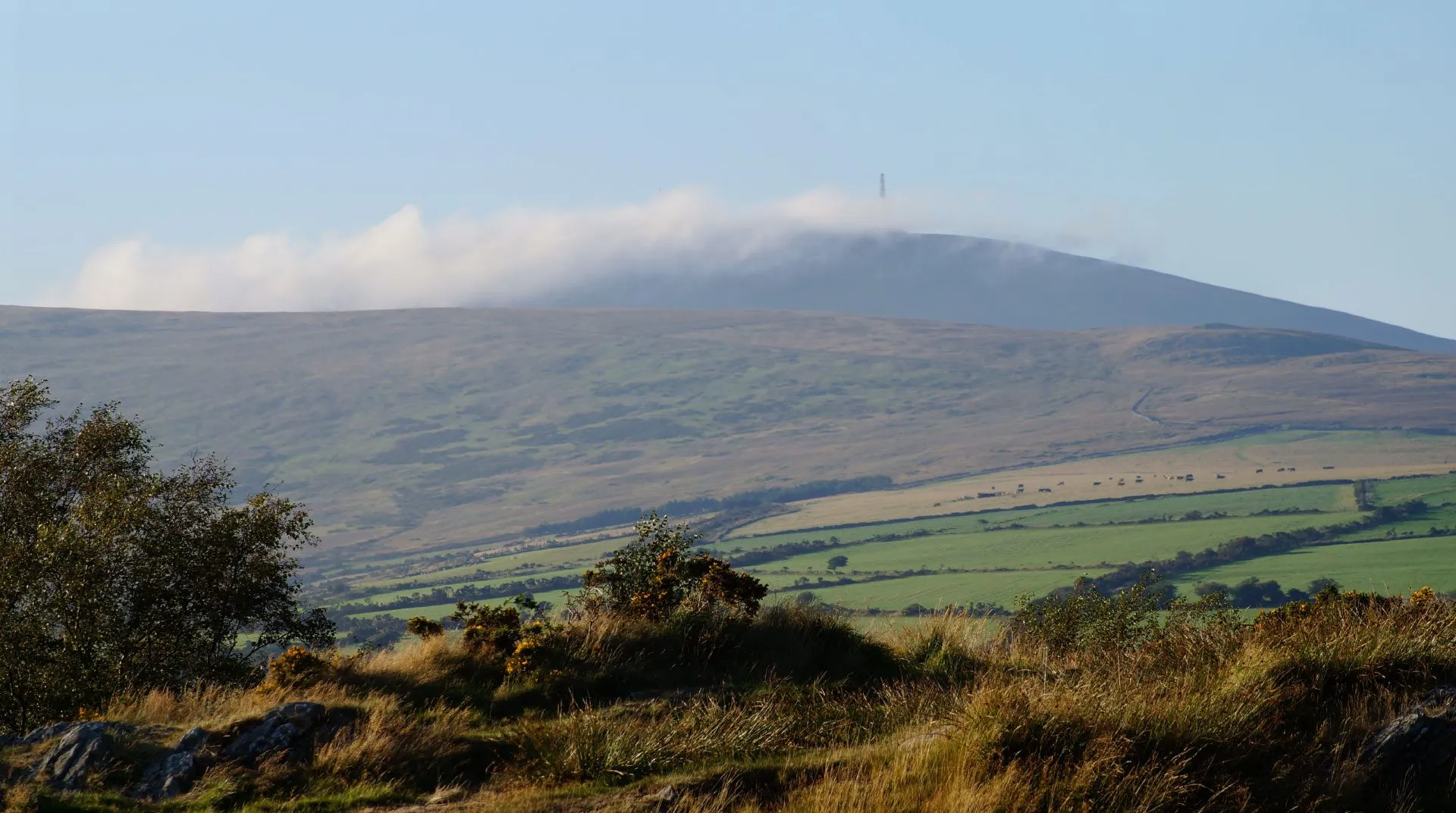 Snaefell's summit and mast wrapped in a band of white cloud above brown moorland and green fields, under an otherwise clear blue sky