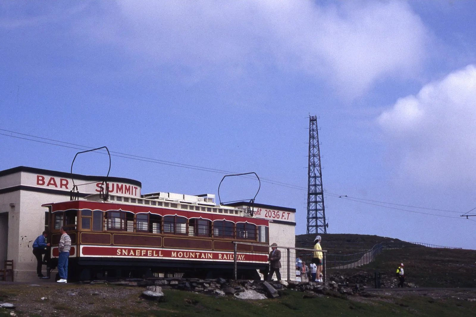 A Snaefell Mountain Railway tram standing at the summit station building, with a radio mast behind and blue sky above