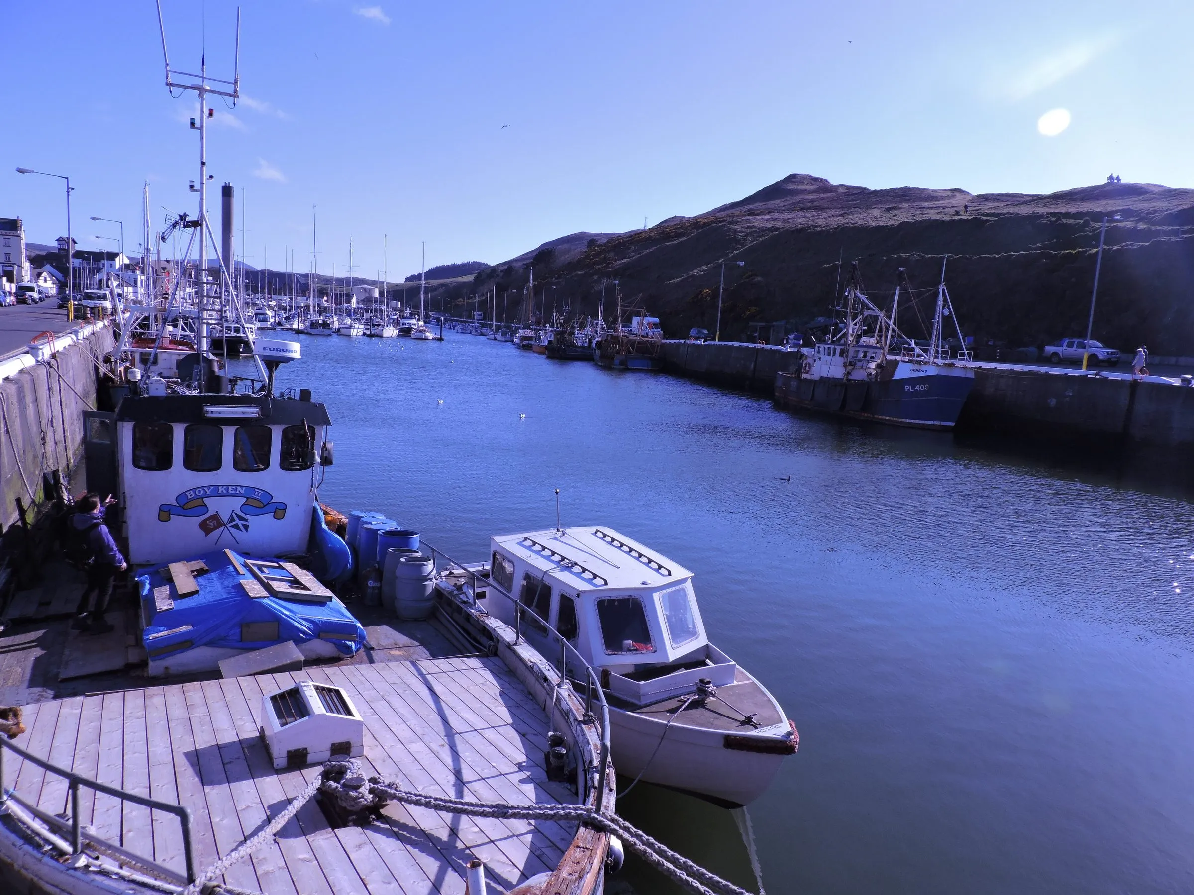 Fishing boats moored in Peel harbour on a sunny day, with the marina stretching back towards the green hills behind the town