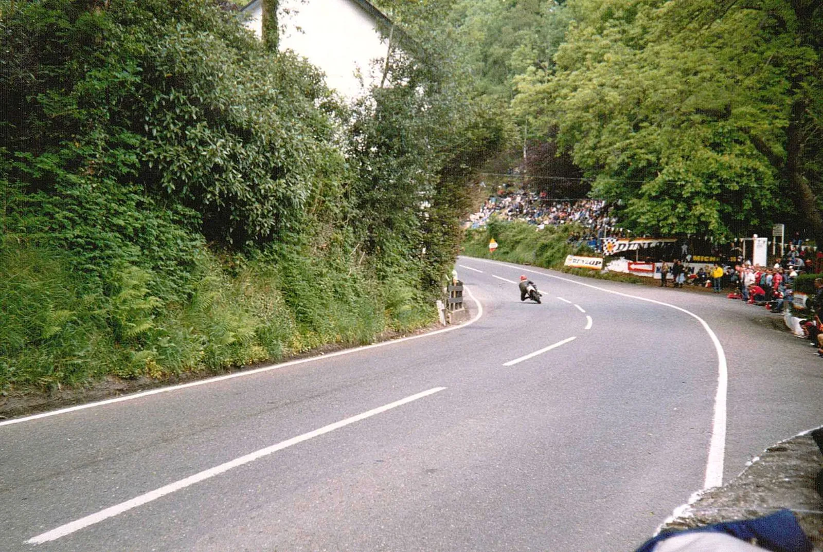 A rider leaning into a curve at Glen Helen during the TT, between a green hedge and a crowd of spectators on the bank opposite