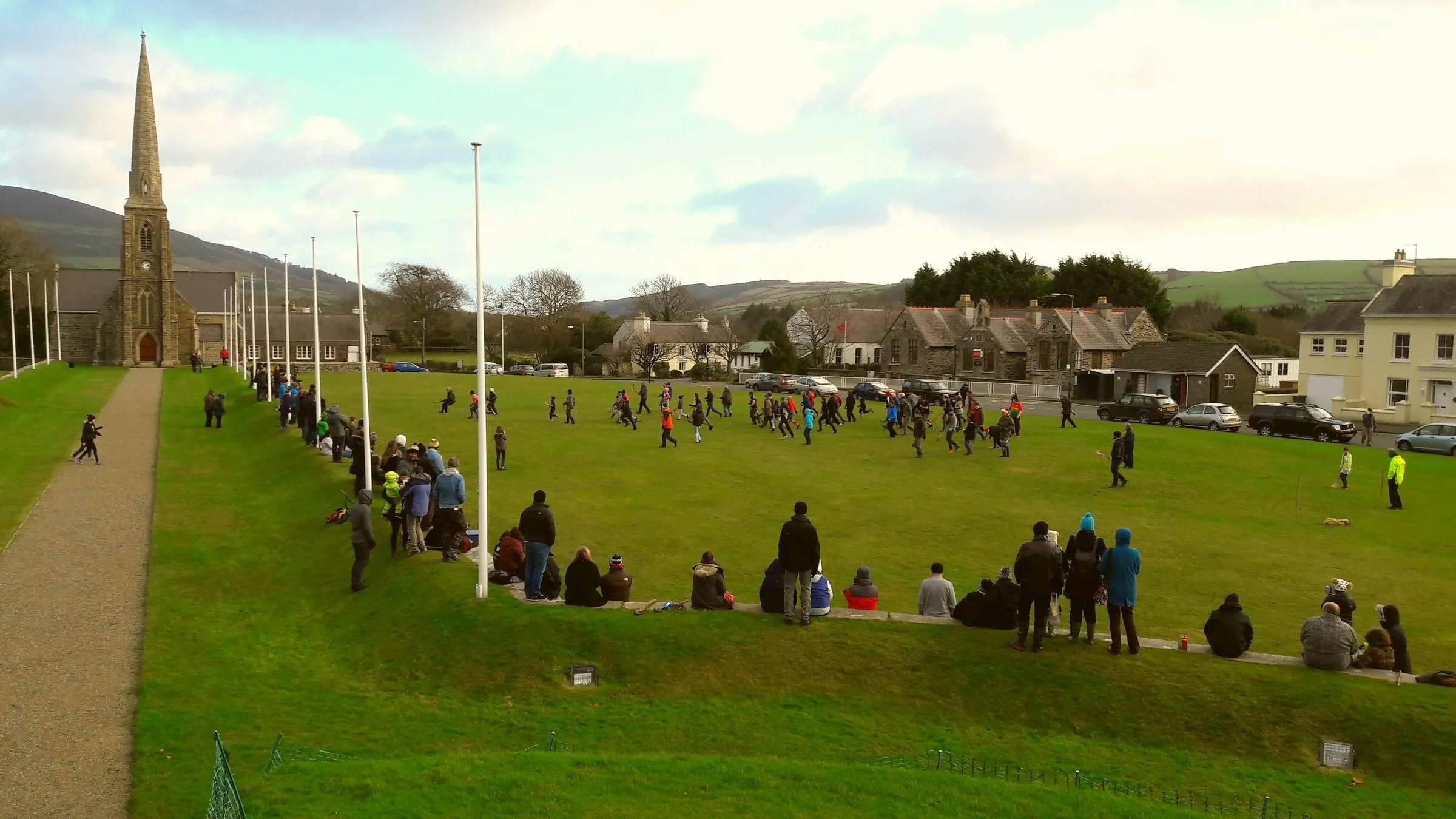 A crowd playing the St Stephen's Day cammag match on the green at St John's, watched from grass banks beside the processional path, with the church spire behind