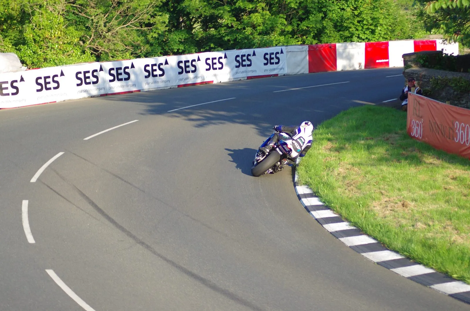 A superbike rider leaning hard through the left-hander at Sulby Bridge, between red and white barriers and a grass verge