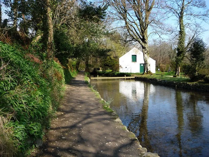 A path beside the still millpond at Silverdale Glen, with a white cottage under the trees on the far bank