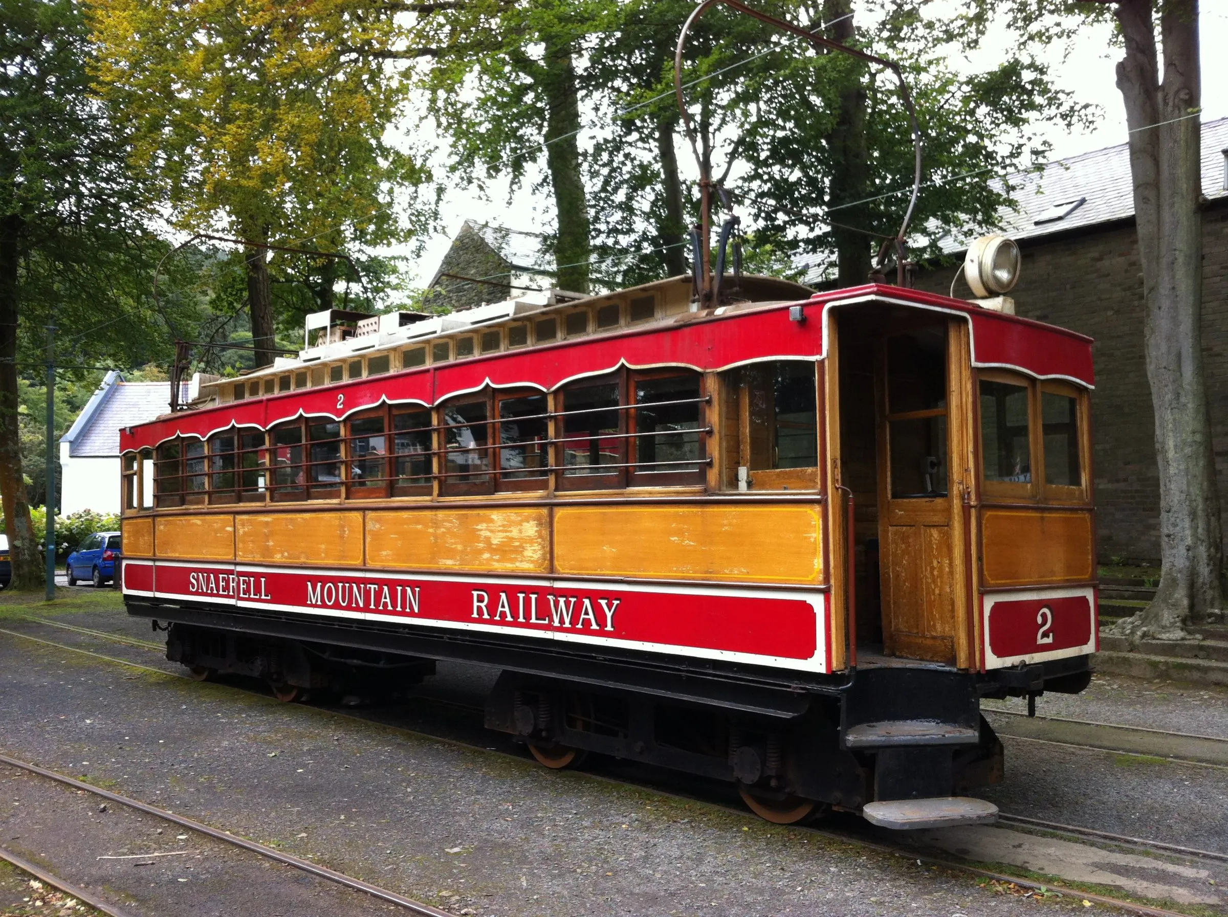 A red and varnished-wood Snaefell Mountain Railway tram, number 2, waiting on the tracks at Laxey under tall trees
